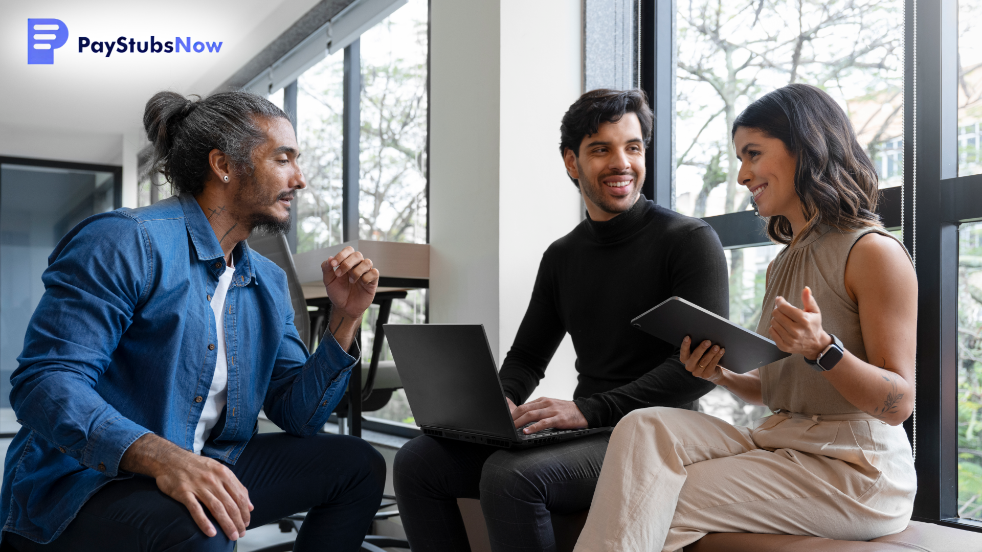 Three people sit together in an office engaging in conversation while one person types on a laptop