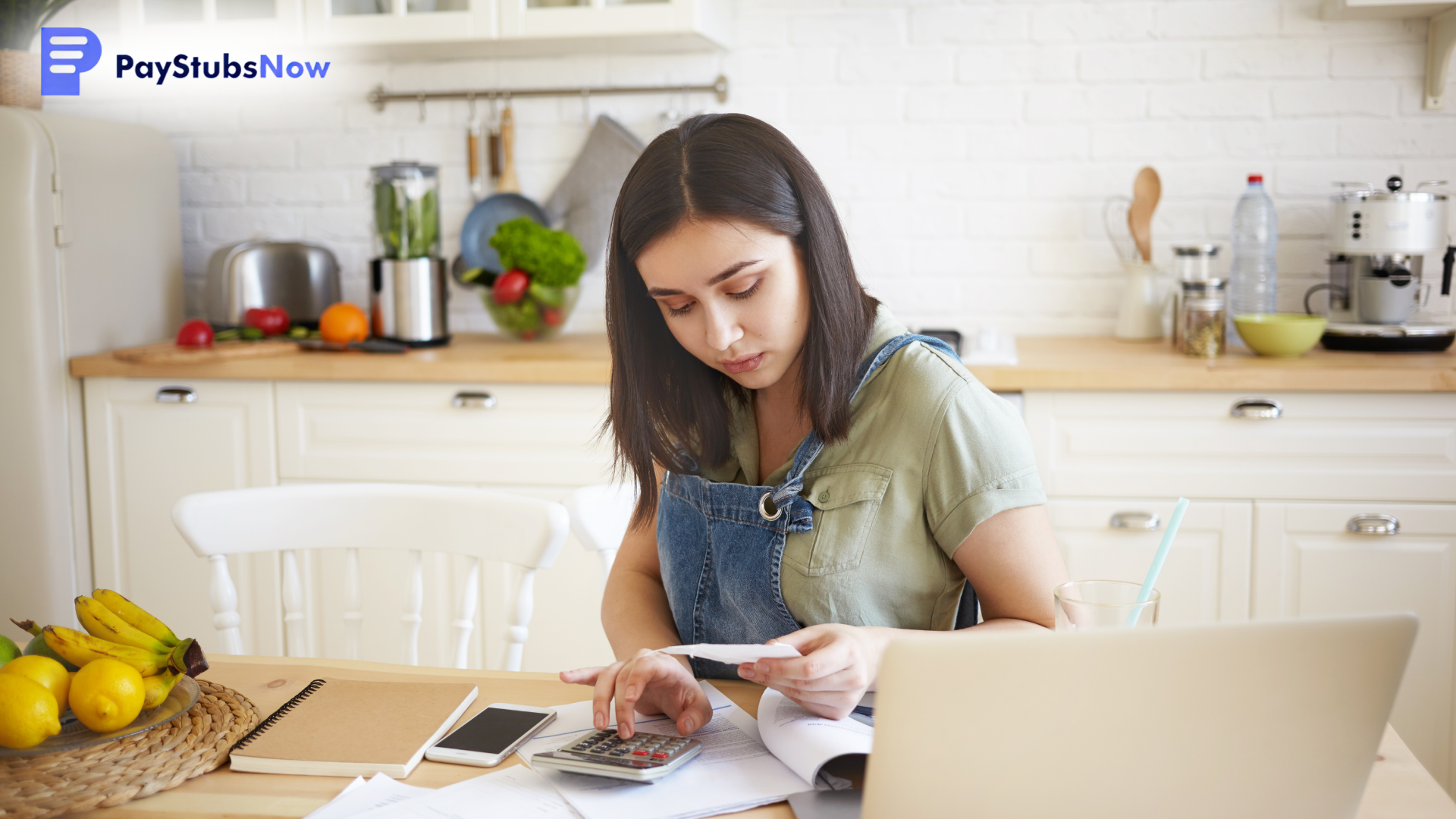 A woman sitting at a wooden kitchen table, wearing overalls, while using a calculator and looking at a receipt.