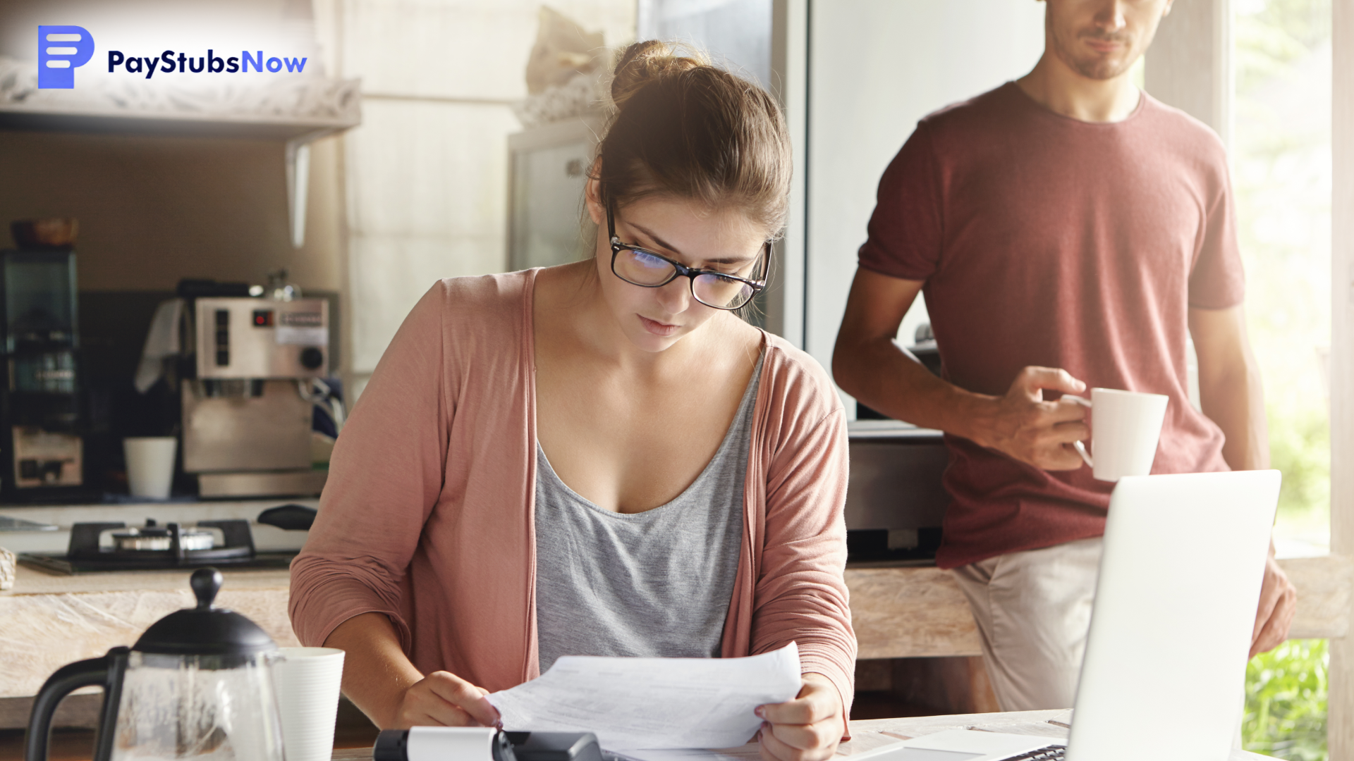 A woman wearing glasses reviews a document at a table while a man stands behind her holding a coffee mug.