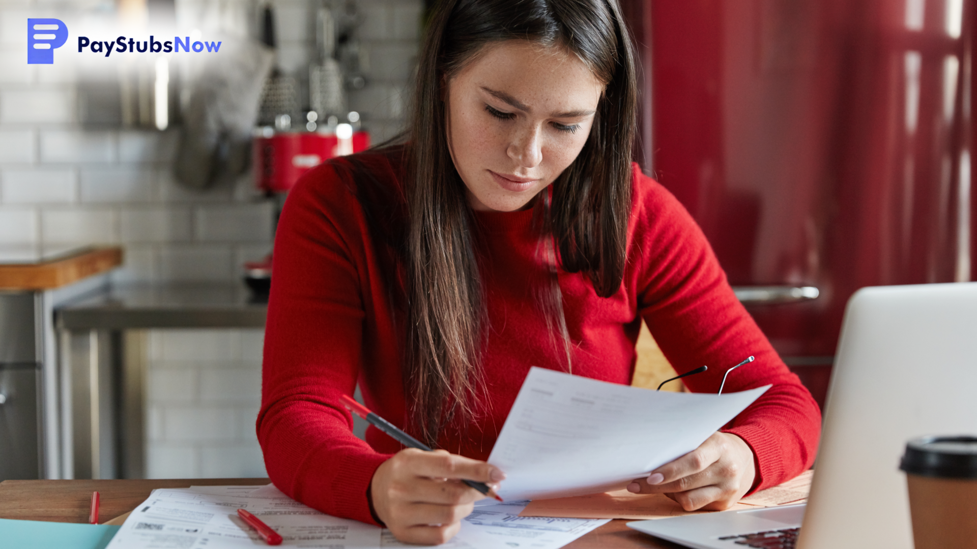 A woman sits at a table with papers and a laptop, reading a document and holding a pen.