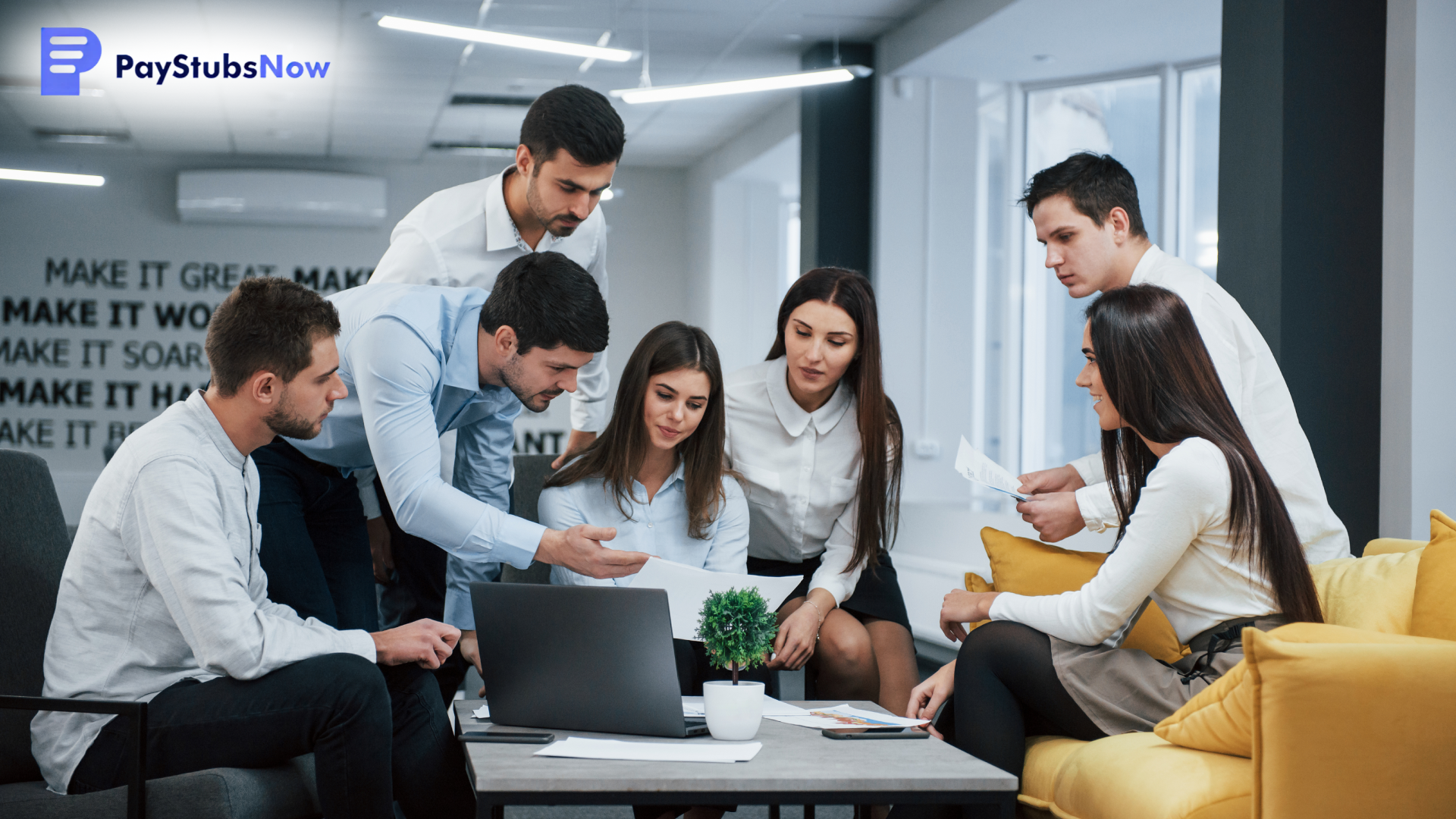 A group of employees, sitting and standing around a small table with a laptop, look at a document together in a modern office.