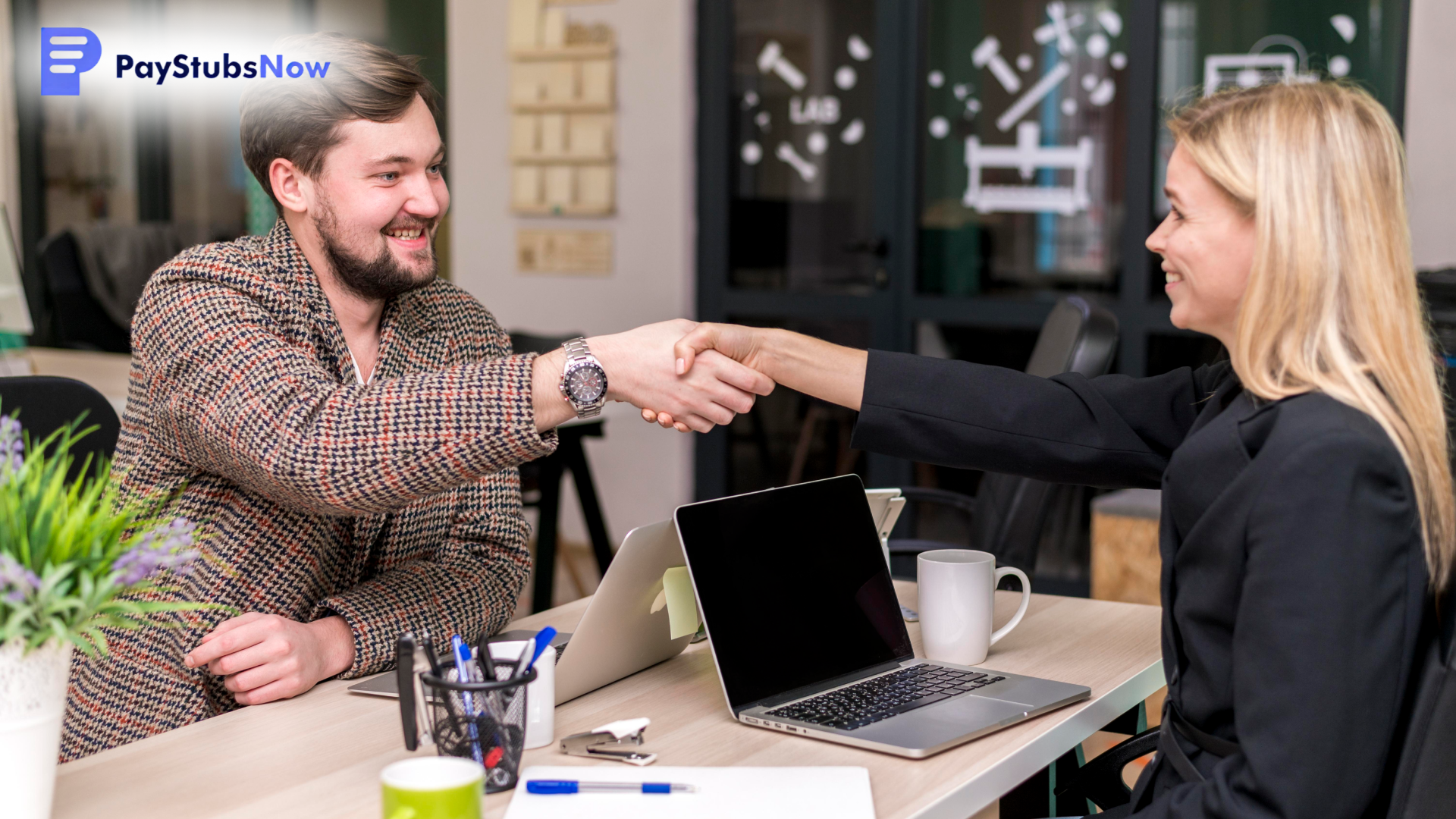 A man and a woman are seated at a desk with a laptop and shaking hands, smiling at each other.