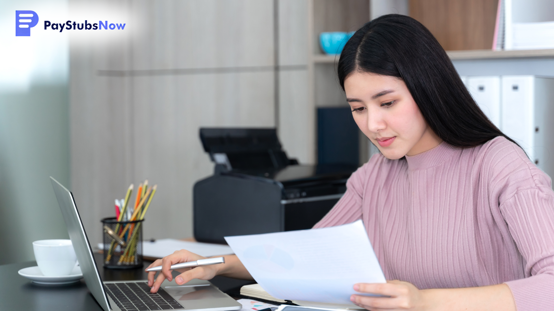 A woman sits at a desk using a laptop and reviewing a document.